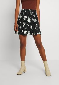 Jupe fleurie noire avec une taille froncée, ornée de fleurs blanches et roses et de feuilles vertes, associée à des bottines beiges à talons.