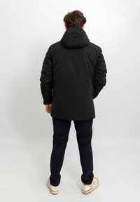 Giacca imbottita nera con cappuccio, caratterizzata da una finitura liscia e opaca. Indossata con pantaloni blu navy e scarpe sportive bianche, vista da dietro.