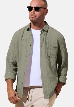 Mann trägt texturiertes olivgrünes Hemd mit Knöpfen über weißem T-Shirt, beige Hose, Sonnenbrille, eine Hand in der Hosentasche, vor einfarbigem Hintergrund.