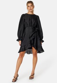 Robe noire portefeuille avec manches bouffantes, ourlet volanté et tissu texturé. Dispose d'une ceinture à nouer à la taille pour un ajustement réglable.