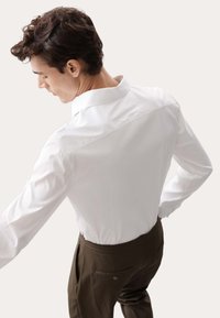 Weißes Langarmhemd mit strukturiertem Kragen, kombiniert mit dunkelbraunen Hosen, die eine Gesäßtasche und dezente Knopfdetails aufweisen.