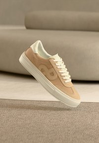 Μπεζ και λευκό αθλητικό παπούτσι με κορδόνια, ανυψωμένο πάνω από υφή μοκέτας, μπεζ καναπές σε φόντο με μαλακή εστίαση.