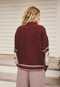 Bordeauxroter Strickcardigan mit gemusterten Ärmeln mit floralen Details, gerippter Textur und entspannter Passform, kombiniert mit Vichy-Karohose.