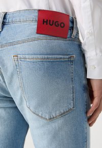 Světlé modré džínové kalhoty s červenou koženou visačkou s nápisem "HUGO" na zadní straně. Obsahují detaily šití a klasický design kapes.