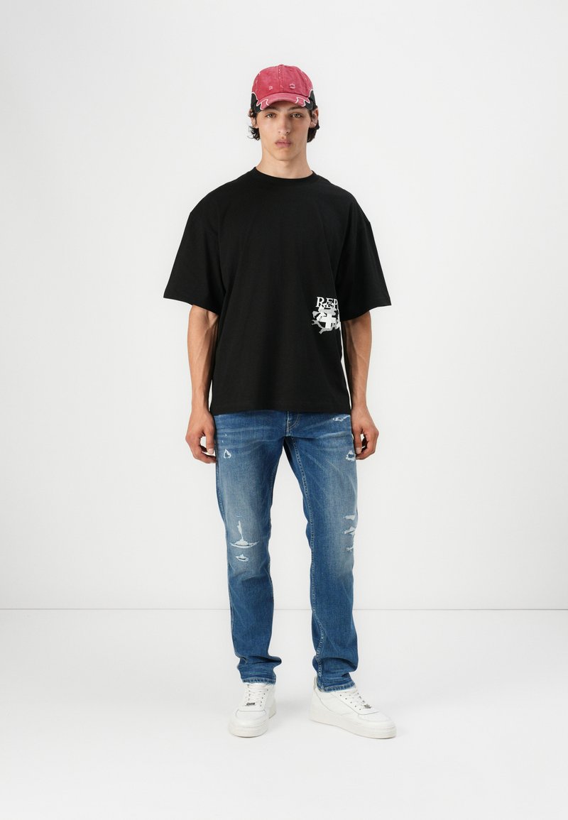 Camiseta negra de gran tamaño con detalle gráfico, combinada con jeans azules desgastados y zapatillas blancas. Una gorra roja añade un contraste de color.