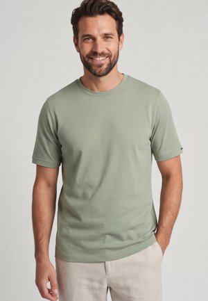 Lächelnder Mann mit dunklem Haar und Bart, der ein hellgrünes kurzärmeliges T-Shirt und helle Hose trägt, vor einem schlichten Hintergrund stehend.