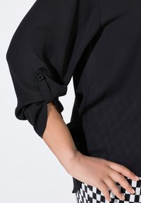 Schwarze Bluse mit einer weichen, glatten Textur, die umgeschlagene Manschetten hat, die mit Knöpfen gesichert sind. Muster sind auf benachbarten karierten Hosen sichtbar.