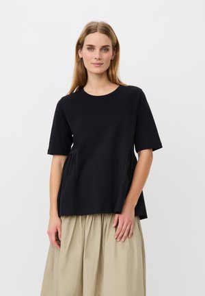Kvinde med langt, glat blondt hår iført en sort kortærmet top og beige plisseret nederdel, stående mod en ensfarvet hvid baggrund.