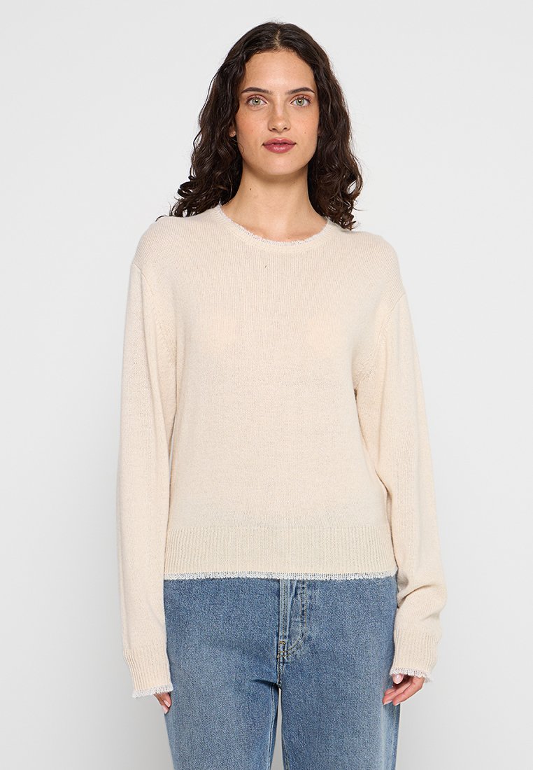 Vrouw met krullend donker haar, gekleed in een crèmekleurige gebreide trui en blauwe jeans, staand tegen een lichte achtergrond.