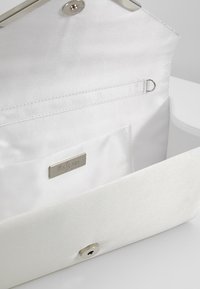Pochette en satin blanc avec une finition lisse, fermeture à pression et petite plaque logo à l'intérieur. L'intérieur comprend une poche plate et des éléments métalliques argentés.