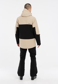 Beige en zwarte waterdichte skijas met een capuchon, met een kleurblokken ontwerp, losse pasvorm en elastische manchetten. Zwarte skibroek maakt de outfit compleet.