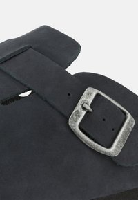 Schwarze Wildleder-Sandale mit einem breiten Riemen, silberner metallischer Schnalle und Ausschnittdesign. Weiche Textur mit einer flachen, robusten Sohle.