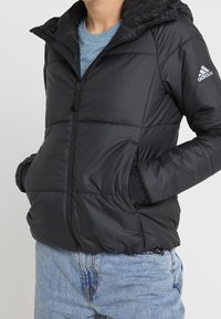 Sort pufferjakke med hætte, der har en frontlynlås, elastiske manchetter og et lille logo på højre ærme. Dennes iført over en blå skjorte.