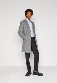 Grahamfarbener Wollmantel mit Reverskragen, zwei Knöpfen und seitlichen Taschen, kombiniert mit einem hellblauen Hemd und schwarzen Hosen, abgerundet durch schwarze Schuhe.