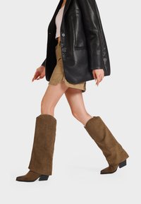 Stivali al ginocchio in suede marrone con punta quadrata e tacco a blocco. Abbinati a un blazer in pelle nera e pantaloni corti kaki.