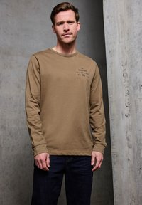 Hnědý bavlněný tričko s dlouhým rukávem a kulatým výstřihem, s potiskem na levé straně hrudi, v kombinaci s tmavě modrými džínami.