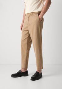 Pantalons en coton khaki avec une coupe droite, des poches latérales et une taille à boutons. Portés avec des mocassins noirs avec un accent doré.