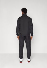 Black tracksuit with elastic cuffs and waistband, smooth material, minimal detailing, worn with white and red athletic shoes.