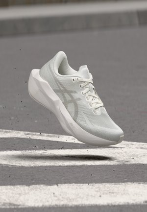 Chaussure de course blanche avec semelle épaisse et dessus en mesh suspendue au-dessus d'une route asphaltée avec des marquages blancs et de petits débris.