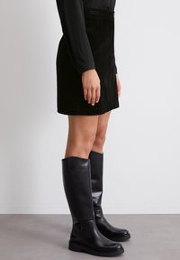 Jupe en velours côtelé noir au design droit, fermeture par bouton, associée à des bottes en cuir noir brillant à talon épais.