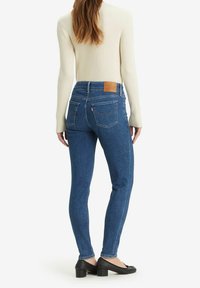 Jeans ajustados de denim de color azul con cintura alta, bolsillos traseros y una etiqueta de cuero marrón en la cintura. La modelo lleva un suéter blanco de canalé.