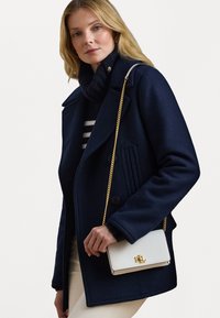 Cappotto in lana blu navy con bottoni doppi e dettagli a righe verticali, abbinato a una borsa bianca con logo dorato e tracolla a catena.