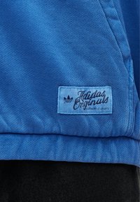 Zila Adidas Originals jaka ar šūtu etiķeti, kurā redzams logotips un virs tā teksta "Authentic Sports", virs melnām biksēm.