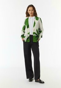Groene en witte cardigan met bladpatroon, knoopsluiting, gedragen over een wit shirt en zwarte broek, gecombineerd met zwarte schoenen.