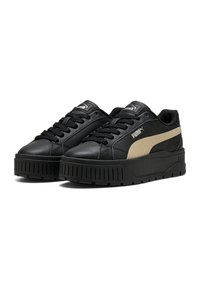 Sneakers neri in materiale sintetico con una suola nera spessa, dotati di una striscia laterale color beige e superficie testurizzata. Lacci neri e linguetta con logo in evidenza.
