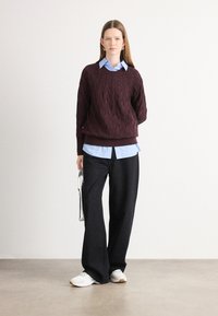 Burgundi kabelio mezgimo megztinis uždėtas ant šviesiai mėlynos marškinėlių su apykakle, derinamas su juodomis plačiomis kelnėmis su subtiliais raštais ir baltomis sneaker'iais.