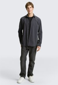 Dunkelgraues Hemd zum Zuschnüren über einem schwarzen T-Shirt, kombiniert mit dunklen Jeans und weißen Turnschuhen. Lässige Passform, glatter Stoff, schlichtes Design.