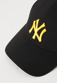 Juodas beisbolo kepurė su išsiuvinėtu geltonu "NY" logotipu. Medžiaga glotni, su matomomis siūlėmis, o snapelis šiek tiek išlenktas.