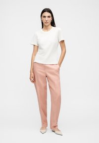 Γυναίκα με μακριά σκοτεινά μαλλιά που φοράει λευκό τοπ με κοντά μανίκια, ανοιχτό ροζ παντελόνια και μπεζ μυτερά παπούτσια, στέκεται με το ένα χέρι στην τσέπη.