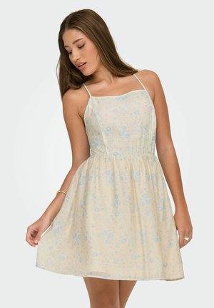 Vrouw in een mouwloze, lichtgele jurk met een blauw bloemmotief, poseert met één hand die de rok vasthoudt terwijl ze naar beneden kijkt.