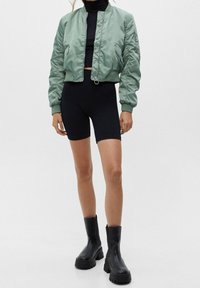 Cropte mintgroene bomberjack met elastische manchetten, zwarte nauwsluitende korte broek en stevige zwarte enkellaarsjes met dikke zool.