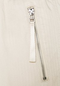 Nahaufnahme eines silbernen Metallreißverschlusses mit einem hellbeigen Stoffzieher auf einem cremefarbenen, strukturierten Stoff.