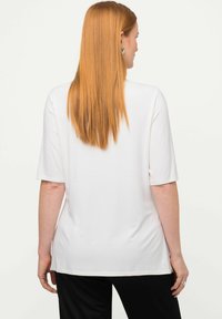 Camiseta blanca de manga corta con un corte holgado, textura suave y dobladillo redondeado, llevada con pantalones negros, mostrada desde la espalda.