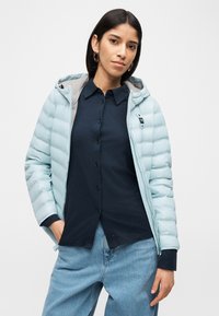 Γυναίκα με σκουρόχρωμα μαλλιά που φορά χρυσούς κρίκους στα αυτιά, ένα ανοιχτό μπλε μπουφάν τύπου puffer πάνω από μια μπλε σκούρα μπλούζα και ανοιχτό μπλε τζιν, στέκεται με το ένα χέρι στην τσέπη.