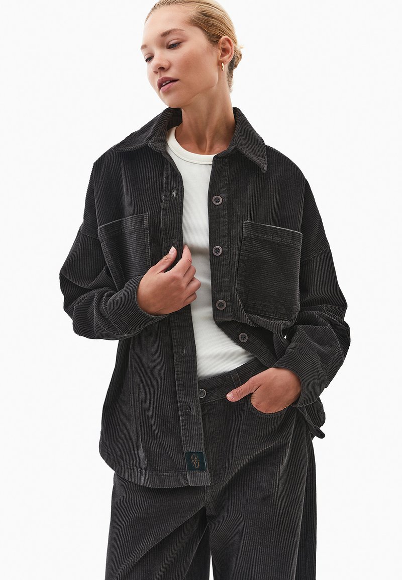 Sort fløjlsk jacket med to forlommer, knaplukning og struktureret stof. Båret over en hvid top med løs pasform og krave.