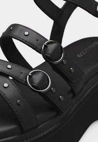 Sandale à plateforme noire avec trois brides en cuir ornées de boucles rondes en argent et de détails cloutés sur fond blanc.