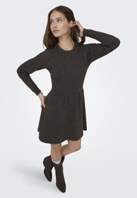 Femme en robe côtelée à manches longues de couleur marron foncé, portant des bottines marron et des chaussettes blanches, posant avec une main sur la hanche et l'autre touchant ses cheveux.