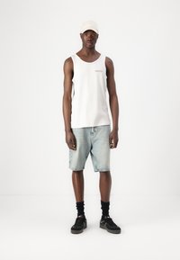 Canottiera grigia chiara con stampa sul petto, pantaloni corti in denim blu chiaro, calze nere e sneakers nere, abbinati a un cappellino beige.