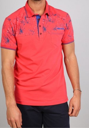 Homme portant un polo rouge avec un motif floral bleu marine sur les épaules et les manches, assorti à un short bleu marine foncé.