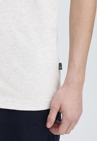 Camisa gris claro hecha de una tela suave, con una etiqueta verde a un lado; una mano toca el dobladillo inferior, con un fondo navy visible.