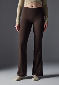 Pantalones marrones acampanados hechos de tejido elástico. Cintura alta y detalle de costura a lo largo de las piernas. El modelo lleva calcetines de color claro.