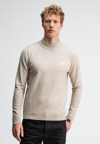 Beige Strickpullover mit hohem Rippkragen und Rippbund. Verfügt über einen kleinen Logo-Patch auf der Brust. Das Material wirkt weich und strukturiert.