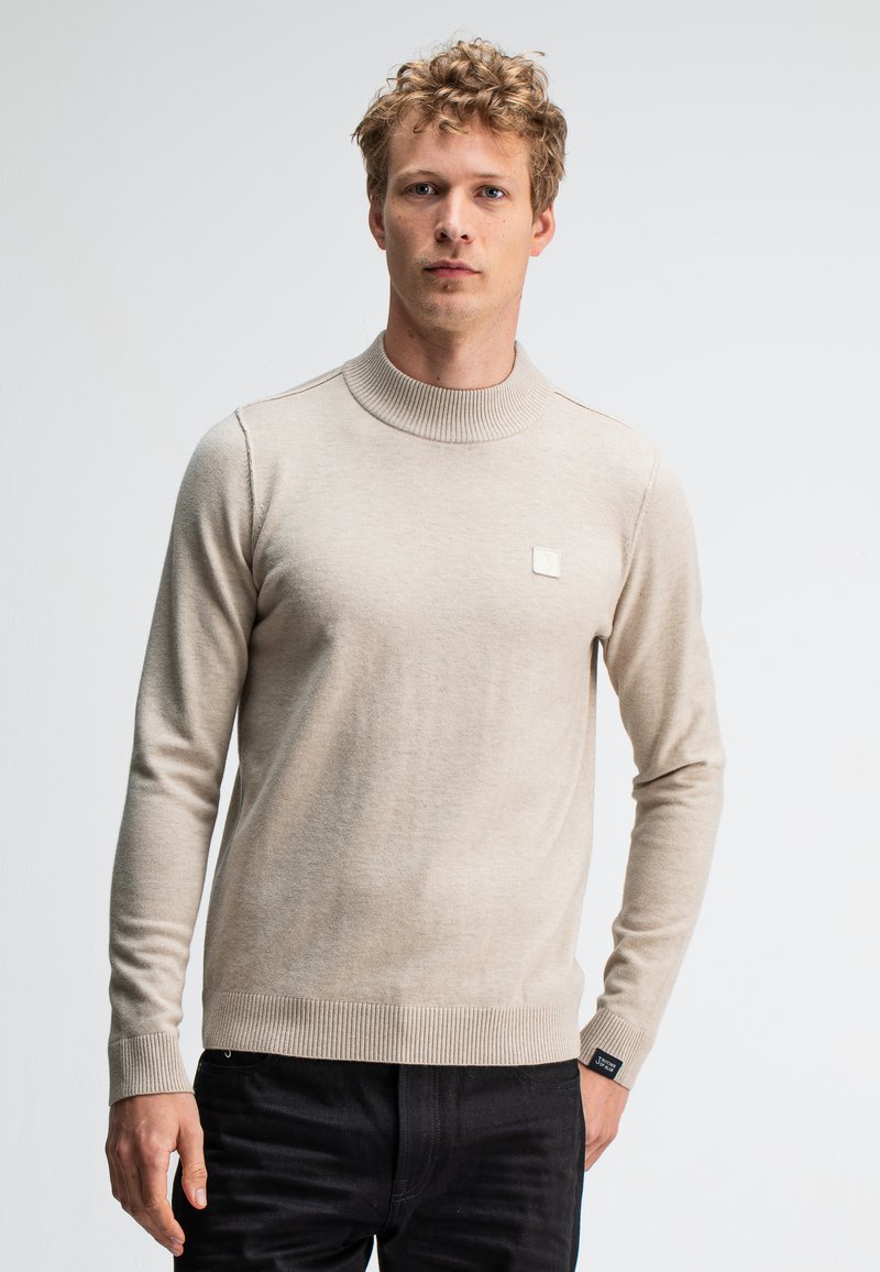 Beige Strickpullover mit hohem Rippkragen und Rippbund. Verfügt über einen kleinen Logo-Patch auf der Brust. Das Material wirkt weich und strukturiert.