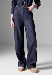 Tumman denim-kankaan korkeavyötäröiset housut, joissa on leveä lahje, etutaskut, nappikiinnitys ja sileä pinta.