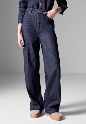 Pantalons taille haute en denim foncé avec une jambe large, dotés de poches avant, d'une fermeture à boutons et d'une texture lisse.