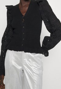 Schwarze Strickjacke mit V-Ausschnitt, fünf Knopfverschluss und Puffärmeln mit Lochmuster. Verfügt über einen gerafften Saum und wird mit glänzenden silbernen Hosen kombiniert.